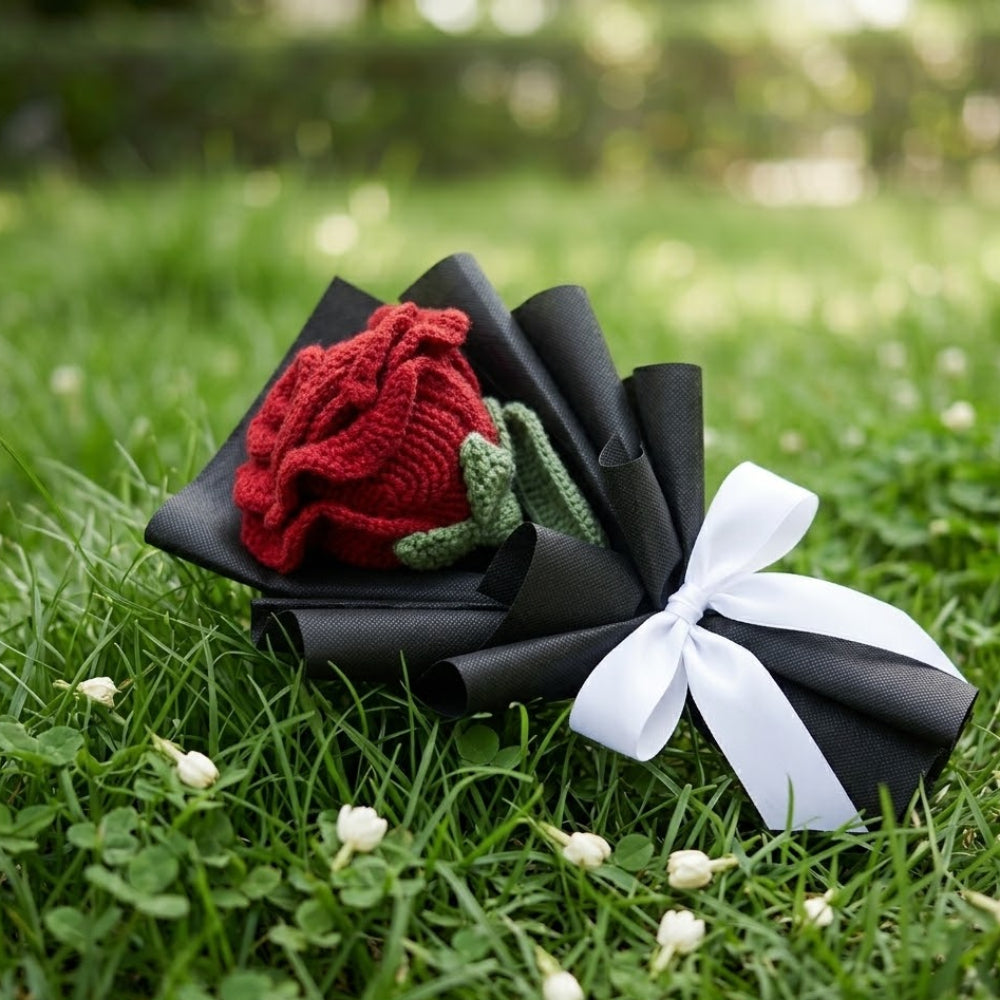 Knitted red rose in a black paper bouquet with a white ribbon on grass
