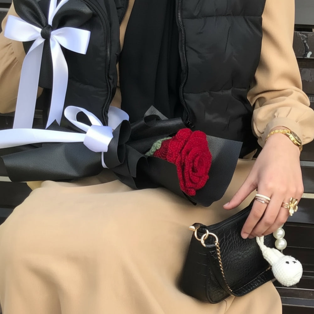 Person sitting on a bench wearing a beige coat, black boots, and holding a small black handbag with a red rose.